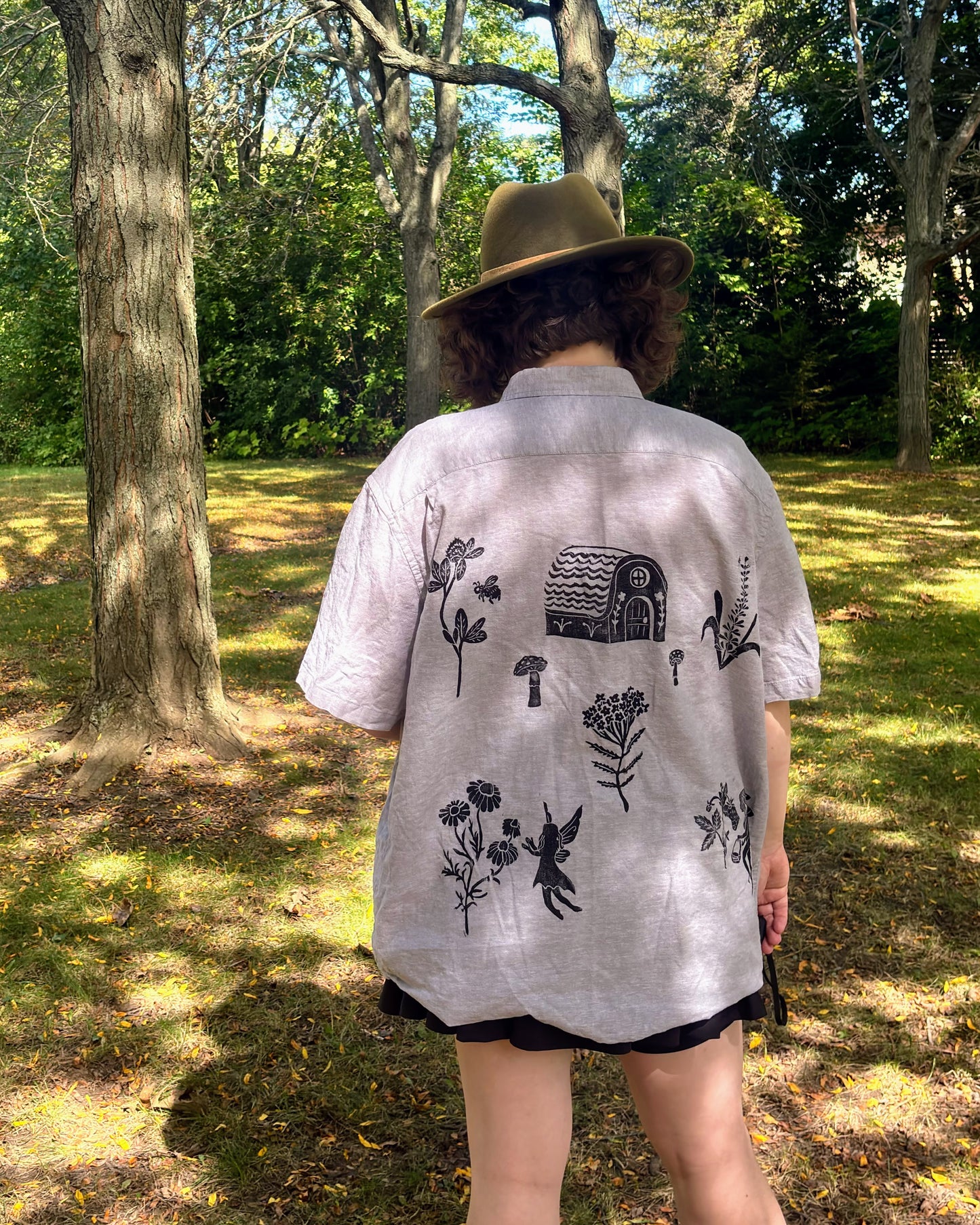 Over the Bridge; grey short sleeve top featuring a bicycle, bridge, and fairy cottage