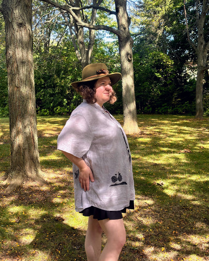 Over the Bridge; grey short sleeve top featuring a bicycle, bridge, and fairy cottage